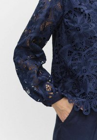 Granatowa koronkowa bluzka z kwiatowymi wzorami i przezroczystymi rękawami, o teksturowanym wykończeniu i marszczonych mankietach.