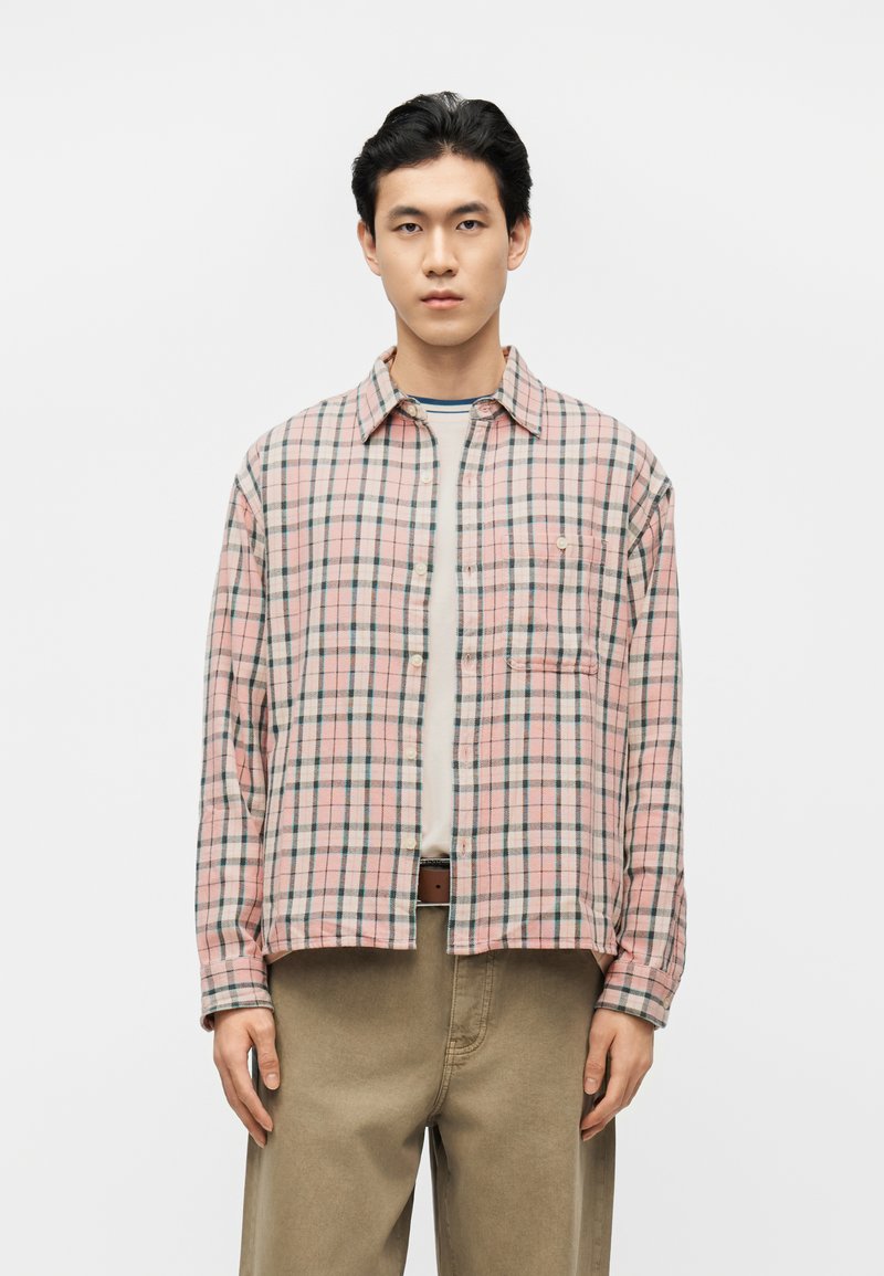 Jeune homme debout, portant une chemise à carreaux rose et verte boutonnée par-dessus un t-shirt blanc, avec un pantalon beige et une ceinture marron, sur un fond blanc uni.