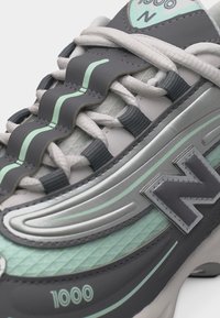 Κοντινή λήψη ενός γκρι και μέντας αθλητικού παπουτσιού New Balance 1000 με λεπτομέρειες στα κορδόνια, υφή στην επάνω πλευρά και εντυπωσιακό λογότυπο "N" στο πλάι.