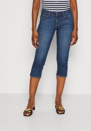 Jean capri en denim bleu foncé avec cinq poches, taille mi-haute et longueur raccourcie, associés à des sandales noires à enfiler avec un accent doré.
