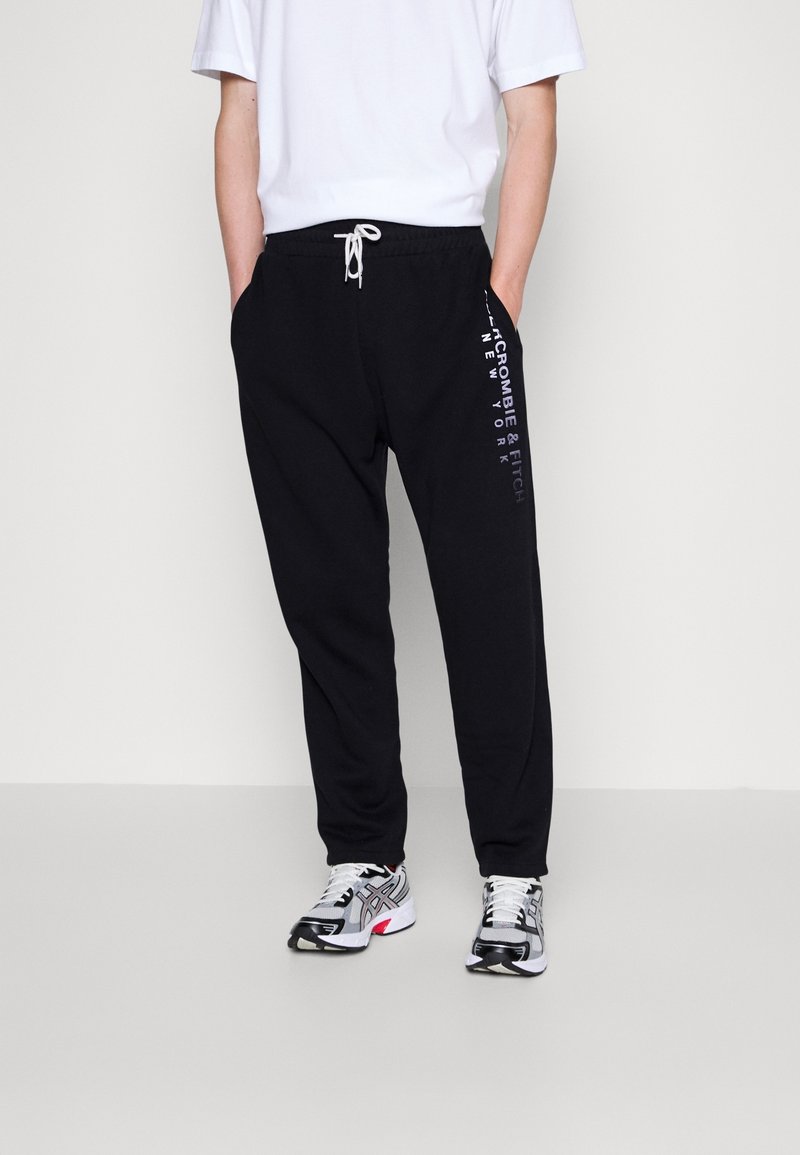 Pantalones de chándal negros con una cintura elástica y cordón, que presentan un estampado de logo blanco en el lado izquierdo. Combinados con una camiseta blanca y zapatillas.