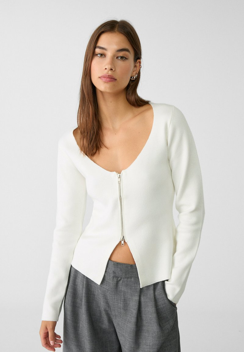 Maglione bianco aderente con maniche lunghe, scollatura a V profonda, zip frontale e orlo asimmetrico. Indossato con pantaloni grigi a vita alta.