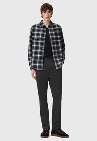 Camicia a scacchi bianca e nera con chiusura a bottoni, indossata sopra un maglione nero, abbinata a pantaloni scuri e scarpe nere con suola marrone.