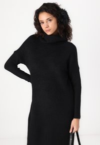 Čierne pletené šaty s vysokým golierom, s rebrovanou textúrou, dlhými rukávmi a voľným strihom. Modelka drží čiernu kabelku s elegantným dizajnom.
