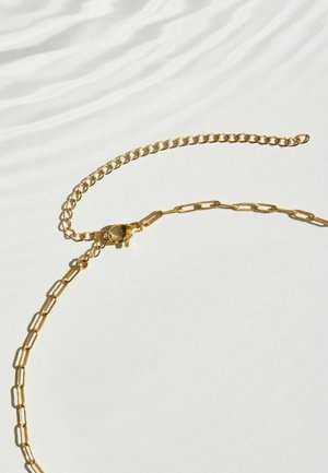 Goldene Halskette mit länglichen Gliedern und einer verstellbaren kleineren Gliederkette auf einer weißen Oberfläche mit weichen Schatten.