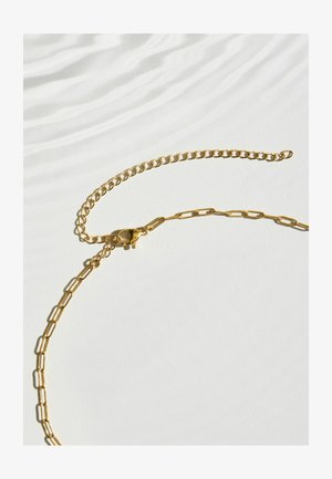 Goldene Halskette mit länglichen Gliedern und einer verstellbaren kleineren Gliederkette auf einer weißen Oberfläche mit weichen Schatten.