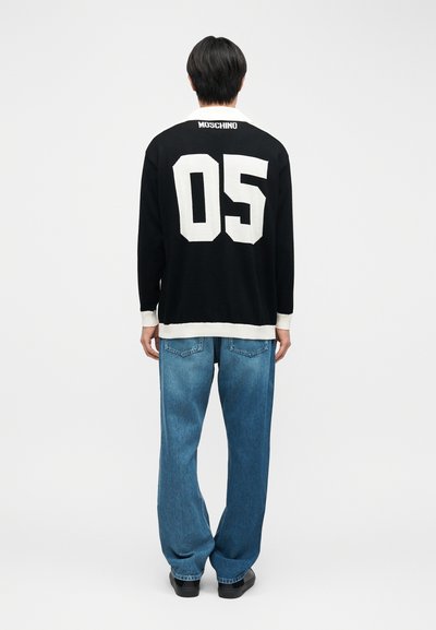 Άτομο που φοράει μαύρο πουλόβερ με λευκό κείμενο "05" και "MOSCHINO" στην πλάτη, μπλε τζιν και μαύρα παπούτσια, στέκεται μπροστά σε λευκό φόντο.