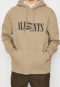 Beżowa bluza z kapturem, kieszenią typu kangur, sznurkiem w kapturze i dużym czarnym napisem z przodu. Miękki materiał o luźnym kroju.