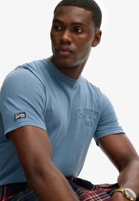 Camiseta de cuello redondo de color azul claro, hecha de algodón, con un logotipo texturizado en el pecho y una etiqueta en la manga. Se lleva con pantalones cortos de cuadro.