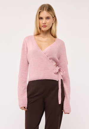 Giovane donna con capelli biondi lisci indossa un maglione rosa incrociato annodato sul fianco e pantaloni marrone scuro su uno sfondo chiaro e semplice.