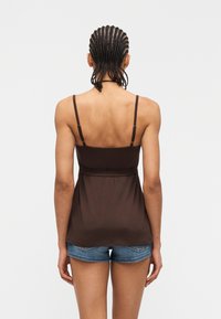 Γυναίκα με πλεγμένα μαλλιά, φοράει καφέ αμάνικη μπλούζα και μπλε τζιν σορτς, στέκεται με την πλάτη προς τον θεατή μπροστά σε απλό φόντο.