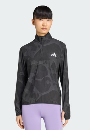 Frau mit schwarzen, glatten Haaren, die ein schwarzes, gemustertes Sportoberteil mit Reißverschluss und Logo sowie hellviolette Leggings trägt, steht vor einem schlichten Hintergrund.