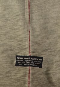 Tessuto beige con linea centrale di cucitura rossa e un'etichetta rettangolare nera con la scritta "ESCAPE FROM / THE UNKNOWN" e testo motivazionale sullo stile.