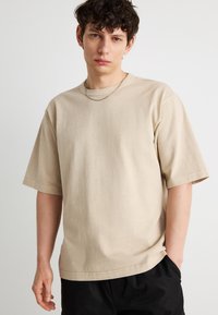 Νέος άνδρας με κοντά σγουρά σκουρόχρωμα μαλλιά, που φοράει μια μπεζ φαρδιά μπλούζα και μαύρο παντελόνι, στέκεται μπροστά σε ένα απλό ανοιχτό φόντο.