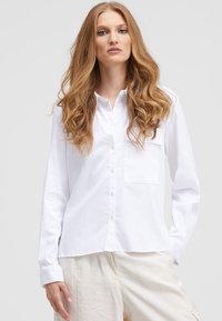 White long-sleeve button-up shirt with a collar, featuring a single chest pocket and a smooth, lightweight fabric. Paired with cream-colored pants.