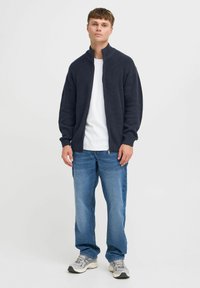 Mørkegrå ribbestrikket jakke med høyt krage, iført over en hvit skjorte, kombinert med lyseblå jeans og grå joggesko.