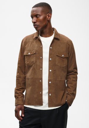 Mann trägt eine braune Wildlederjacke mit Knöpfen über einem weißen Hemd und schwarzen Hosen, schaut nach links mit den Händen in der Nähe der Taschen.