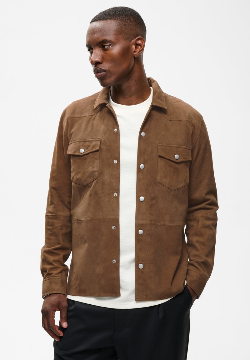 Homme portant une veste en daim marron boutonnée sur une chemise blanche et un pantalon noir, regardant vers sa gauche avec les mains près des poches.