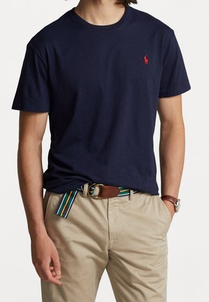 Man draagt een marineblauw T-shirt met korte mouwen en klein rood logo, beige broek, gestreepte stoffen riem en polshorloge, handen in de zakken.