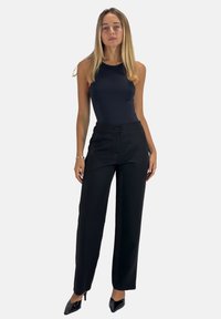 Zwarte aansluitende broek met een hoge taille, zijzakken en een knoopsluiting, gecombineerd met een mouwloos zwart bodysuit en zwarte puntige hakken.