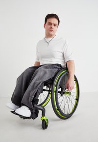 Invalidski s črnim okvirjem in lime zelenimi detajli. Posameznik nosi belo srajco in sive široke hlače, sedi z eno roko na kolesu.