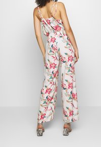 Une combinaison fleurie en tissu doux et léger avec des motifs roses et verts. Elle a des bretelles fines, un dos froncé et des jambes larges avec une coupe ample.