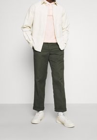 Hombre con zapatillas blancas, pantalones oscuros verde olivo con puños, camiseta polo rosa y una camisa abotonada color crema con las manos en los bolsillos.