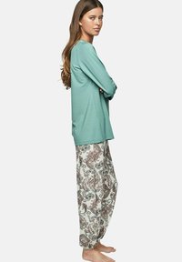 Vrouw die op blote voeten in zijprofiel staat, gekleed in een teal lange mouw top en losse witte broek met paisley patroon tegen een effen achtergrond.