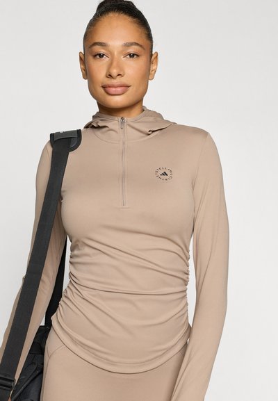 Light brown, long-sleeve athletic top with a half-zip design and hood. Features gathered detail at the waist and a circular logo on the chest.