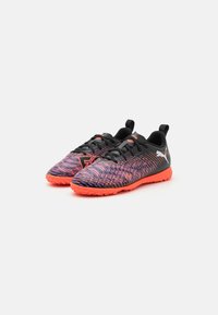 Bottines de football texturées noires et violettes avec une semelle en caoutchouc orange, présentant un design à lacets et un branding proéminent sur le côté.