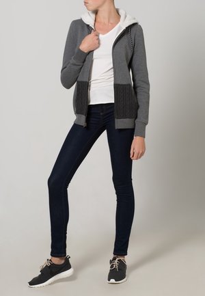 Gray zip-up hoodie with a white fleece-lined hood, cable-knit pockets, paired with dark skinny jeans and black sneakers.