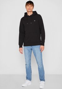 Melna kapuce jaka ar kengura kabatu, aukliņām un emblēmu. Kombinēta ar gaiši zilām džinsa biksēm un baltām sporta kurpēm.