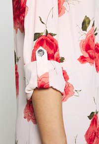 Blouse rose clair avec motif floral rouge, manches courtes roulées et détail en bouton argenté au niveau du poignet. Tissu doux et lisse.