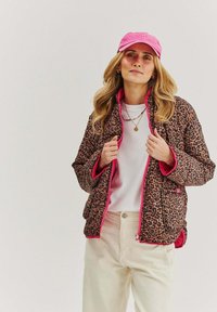 Puchowa kurtka w panterkę w kolorze brązowym i czarnym, wykończona różowym obszyciem. Nosi się ją na białej koszuli, w połączeniu z jasnymi, kremowymi spodniami.