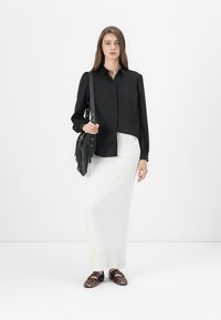 Zwart langemouwen shirt met een knoopsluiting, gecombineerd met een witte textuur maxi rok en schoenen met panterprint, terwijl ze een zwarte franjes tas vasthoudt.