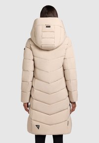 Vrouw in een lange beige pufferjas met een grote capuchon, gezien vanaf de achterkant tegen een effen achtergrond.