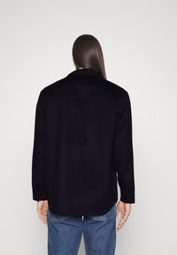 Black, textured overshirt with a collar, button cuffs, and a curved hem. Worn over light blue jeans, viewed from the back against a plain background.
