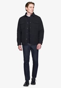 Sort skjortejakke med to brystlommer, båret over en mørk sweater og parret med mørke jeans og sorte sko, sat mod en hvid baggrund.