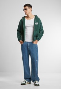 Sudadera verde con cremallera sobre una camiseta blanca sin mangas, combinada con jeans azules holgados y zapatillas verdes y blancas. Se llevan gafas de sol claras.