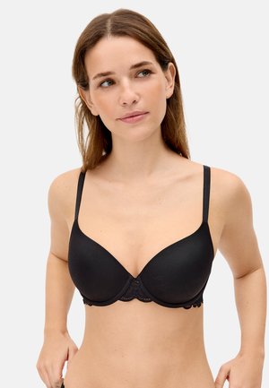 Mujer de piel clara y cabello castaño, con un sujetador acolchado negro de tirantes finos y encaje, mirando ligeramente hacia un lado.