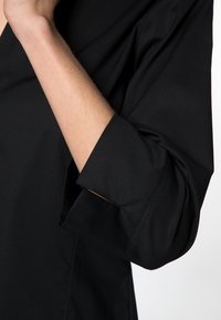 Chemise noire à col en V avec des manches trois-quarts. Présente un tissu lisse, une texture subtile et des poignets soigneusement finis.