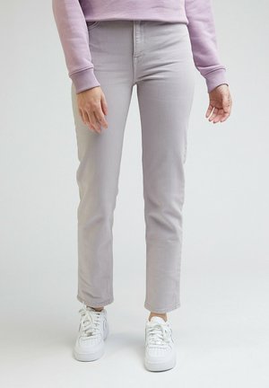 Personne portant un pantalon gris clair à jambes droites, des baskets blanches et un haut à manches longues violet clair, debout devant un fond uni.