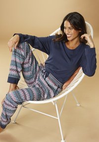 Marinblå långärmad topp i kombination med mönstrade joggerbyxor med rosa, blå och grå zigzag-motiv. Modellen sitter i en vit stol.