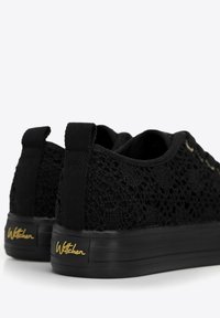 Zapatillas negras con cordones, con parte superior de punto texturizado, suela de goma negra y logotipo dorado en el talón. La vista trasera muestra las lengüetas de agarre.