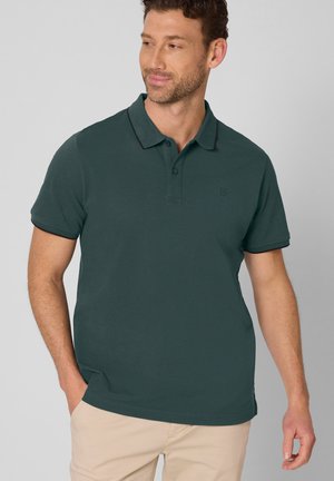 Mężczyzna ubrany w ciemnozieloną koszulkę polo z krótkim rękawem i beżowe spodnie, stojący z jedną ręką w kieszeni, patrzący w bok, neutralne tło.