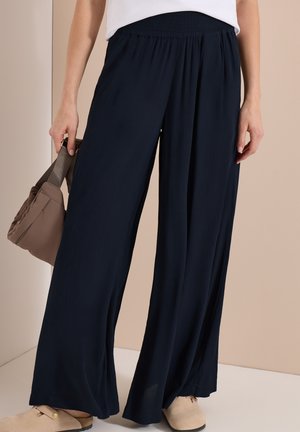 Vrouw draagt losse zwarte broek en beige instapschoenen, houdt een taupekleurige handtas vast tegen een neutrale achtergrond.