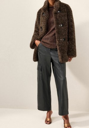 Osoba ubrana w brązowy, futrzany płaszcz na guziki, metaliczny, brązowy sweter dziany, czarne skórzane spodnie o szerokich nogawkach z kieszenią cargo oraz brązowe sandały na obcasie z odkrytymi palcami.