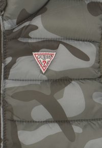 Giubbotto imbottito camouflage in toni di grigio; presenta un patch logo triangolare rosso e bianco "GUESS"; texture trapuntata, chiusura con zip.