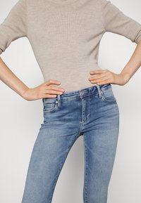 Vaqueros azul claro con un diseño ajustado, estilo de cinco bolsillos, botón de metal y un sutil detalle de desgaste en las piernas. Combinados con un top de punto beige.