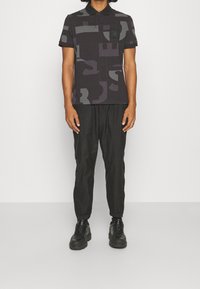 Polo noir avec un motif géométrique en gris foncé, doté d'une poche poitrine et de boutons, associé à un pantalon noir ample et des chaussures épaisses.